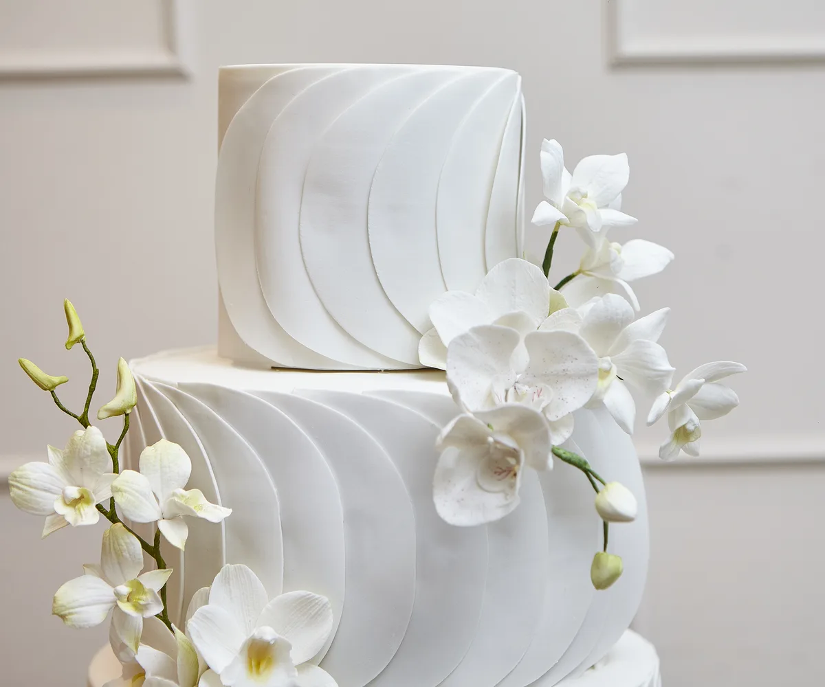 Andares de bolo de casamento branco com orquídeas de açúcar e parede creme ao fundo
