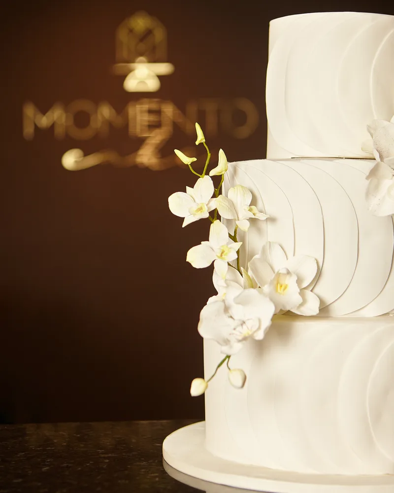 Bolo de casamento plissado com orquídeas ao lado da logomarca dourada Momento Cake