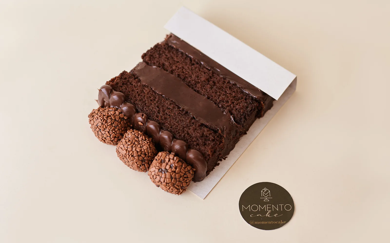 Fatia de bolo de chocolate com ganache e brigadeiros com selo Momento Cake
