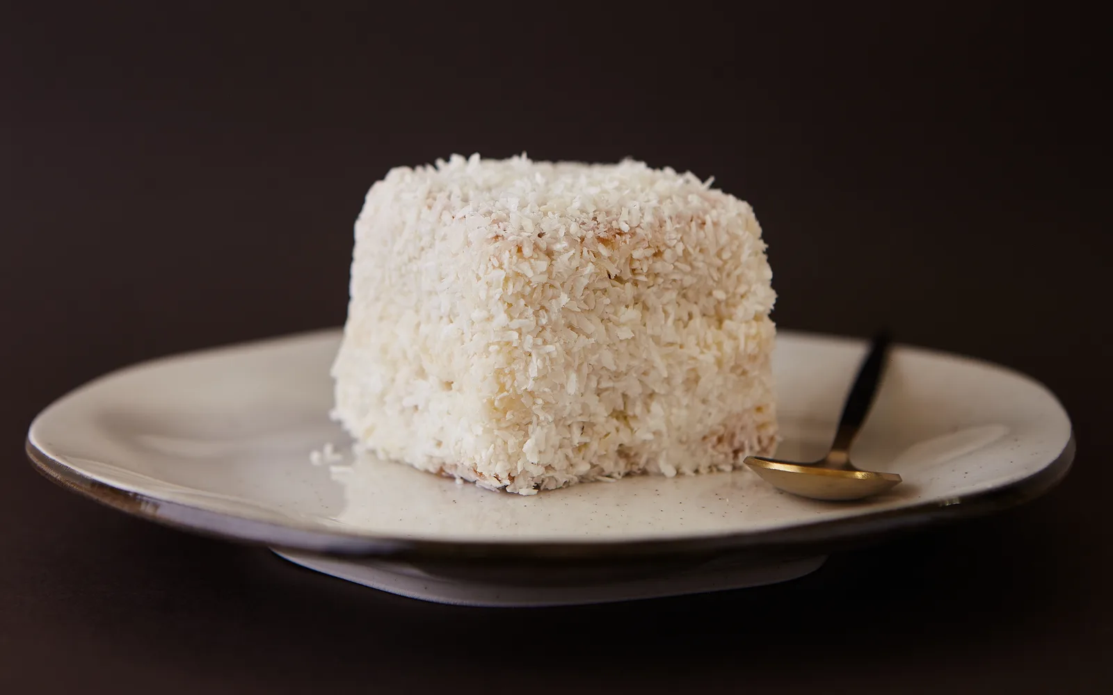Fatia de bolo gelado de coco em prato artesanal com colher dourada sobre fundo escuro