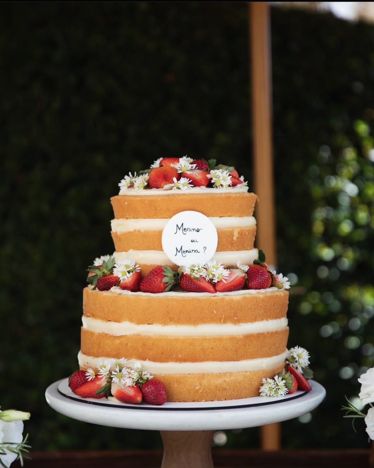 Naked cake de dois andares com camadas de massa branca e recheio cremoso, decorado com morangos frescos, mosquitinho branco e plaquinha de chá revelação Menino ou Menina
